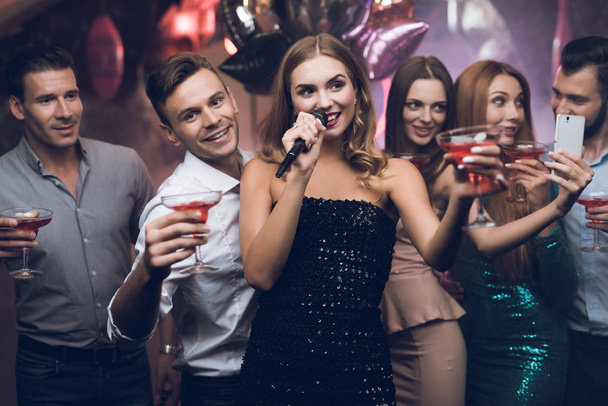 A woman in a black dress is singing songs with her friends at a karaoke club. Her friends have fun on the background. - Φωτογραφία, εικόνα