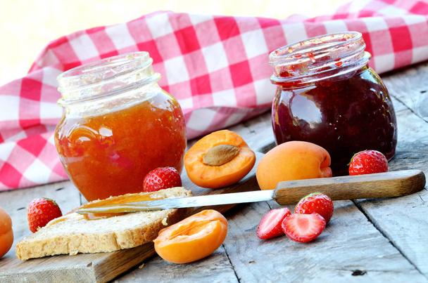 Jar of apricot and strawberry jam, fresh apricots and strawberries, knife and toast bread with butter on wooden table, red checkered tablecloth in background in summer garden - Photo, Image