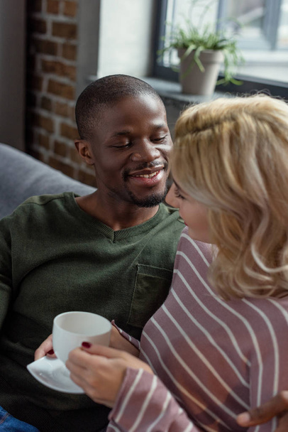 smiling african american man looking at caucasian girlfriend with cup of hot drink in hands while resting on sofa at home - Фото, изображение
