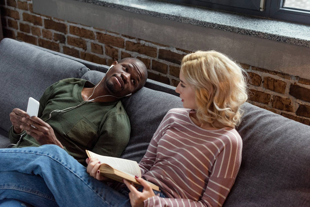 africano americano hombre escuchar música mientras caucásico novia lectura libro en sofá en casa
 - Foto, Imagen