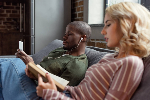 Αφρικανική αμερικανική άνθρωπος ακούγοντας μουσική ενώ Καυκάσιος φίλη διαβάζοντας το βιβλίο στον καναπέ στο σπίτι - Φωτογραφία, εικόνα