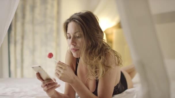 Charming woman using smartphone and licking hard candy on bed - Footage, Video