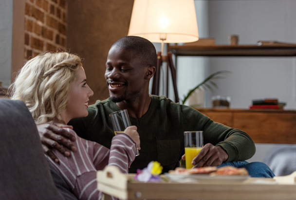 vue latérale du couple interracial souriant avec des verres de jus se regardant à la maison
 - Photo, image