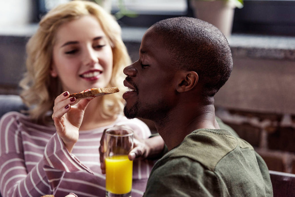 enfoque selectivo de la mujer feliz alimentando novio afroamericano con tostadas durante el desayuno en casa
 - Foto, Imagen