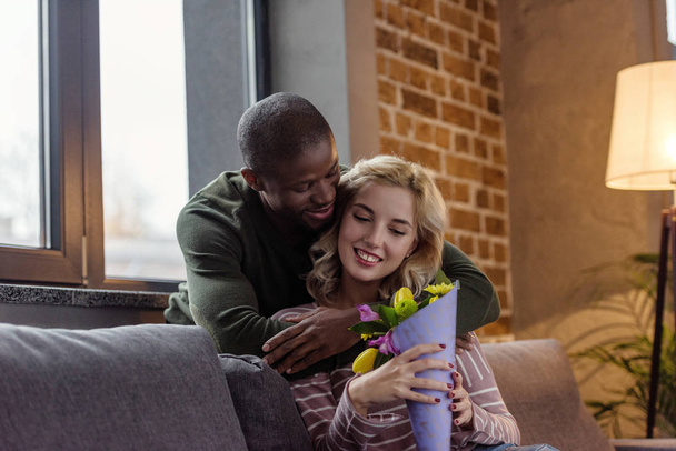 african american man hugging smiling caucasian girlfriend with bouquet of flowers at home - Foto, imagen