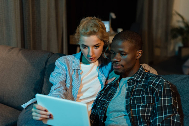 portrait of focused multicultural couple using tablet together while resting on sofa at home - Photo, Image