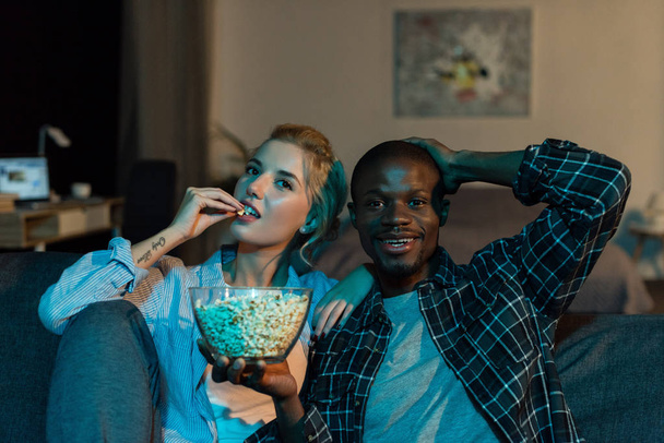 portrait of multiethnic couple eating popcorn and watching film together on sofa at home - Fotografie, Obrázek