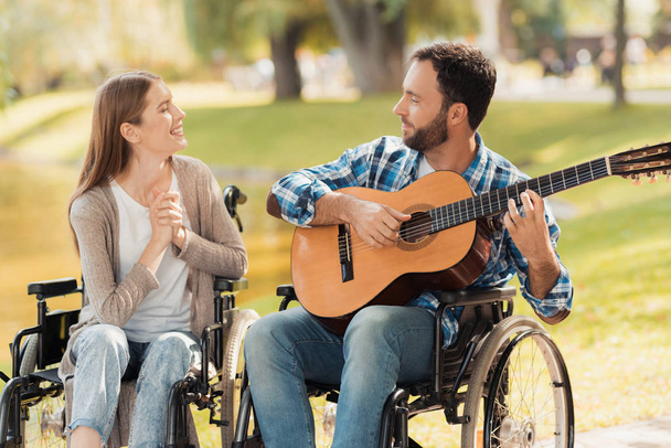 A man and a woman in wheelchairs met in the park. A man is playing the guitar, the woman listens and smiles. - Fotografie, Obrázek
