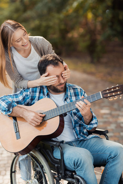 A man on a wheelchair playing guitar in the park. A woman came to him from behind and closed her eyes with her hands. - Foto, imagen