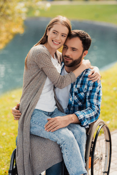 A man is sitting in a wheelchair in a beautiful green park. A woman is sitting on his lap and hugging him. - Foto, Imagem