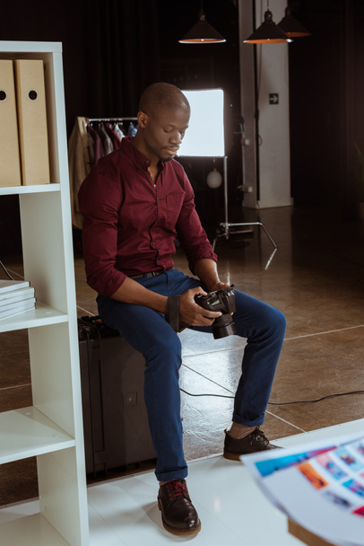 Afrikanischer amerikanischer Fotograf blickt auf Fotokamera in der Hand, während er Fotos im Studio auswählt - Foto, Bild