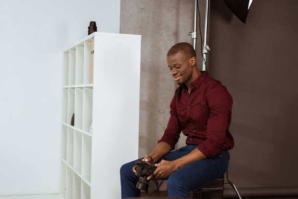 Seitenansicht eines lächelnden afrikanisch-amerikanischen Fotografen, der die Fotokamera in der Hand hält, während er im Studio Fotos auswählt - Foto, Bild
