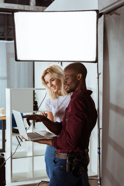 fotógrafo afro-americano e modelo caucasiano escolhendo fotos no laptop juntos durante a sessão fotográfica em estúdio
 - Foto, Imagem