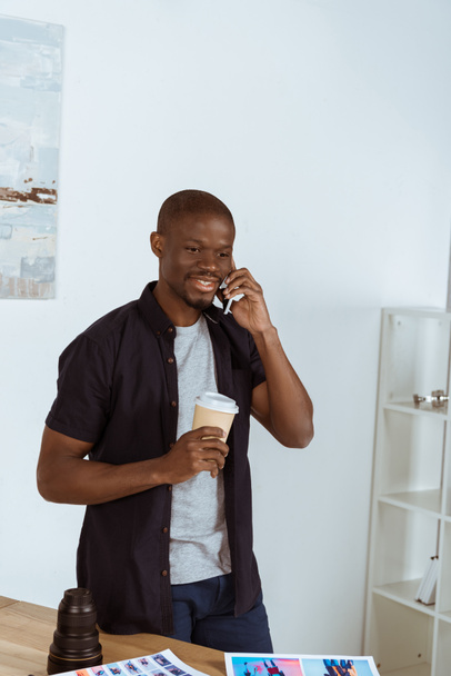 portret uśmiechający się african american człowiek z kawą do przejdź rozmawiać na smartfonie w biurze - Zdjęcie, obraz