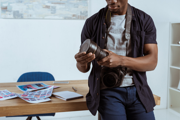 gedeeltelijke weergave van Afro-Amerikaanse fotograaf met fotocamera leunend op de werkplek op kantoor - Foto, afbeelding
