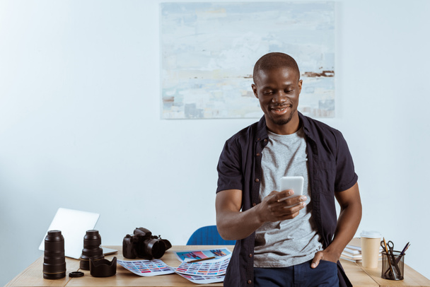 retrato de fotógrafo afro-americano sorridente usando smartphone enquanto se inclina na mesa com equipamentos de fotografia no escritório
 - Foto, Imagem