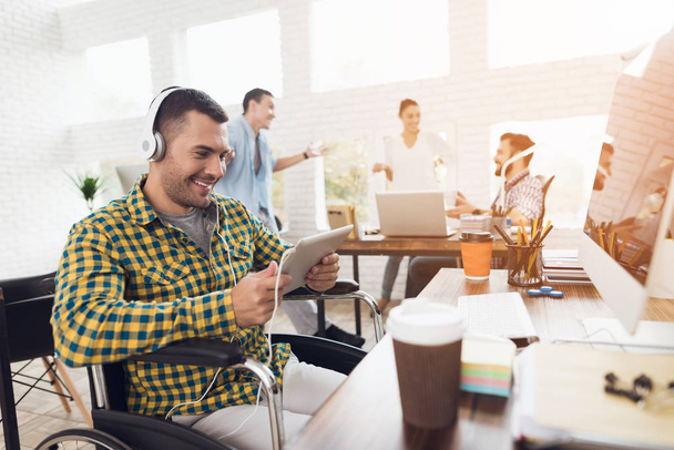 Un hombre en silla de ruedas trabaja con una tableta en una oficina moderna. Escucha música en los auriculares.
. - Foto, imagen