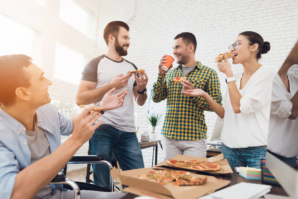 Office workers and man in a wheelchair are eating pizza. They work in a bright and modern office. - Фото, изображение