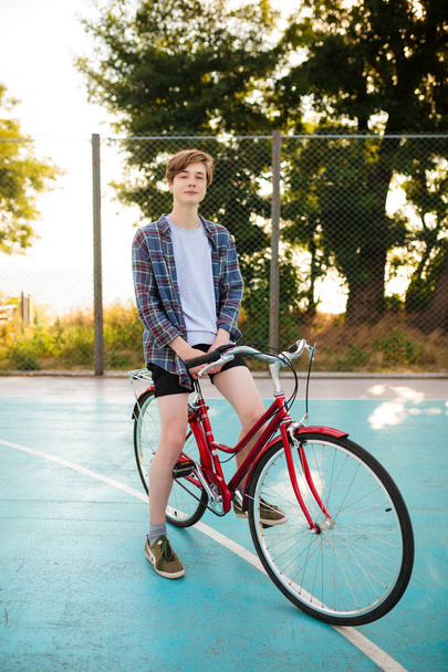 Νεαρός άνδρας με ξανθά μαλλιά στα σορτς και casual πουκάμισο στέκεται με κόκκινο ποδήλατο στο γήπεδο μπάσκετ στο πάρκο. Προσωπογραφία αγοριού που σκεπτικά ψάχνει στην κάμερα ενώ στέκεται με κλασικό ποδήλατο - Φωτογραφία, εικόνα