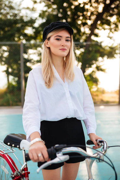 Beautiful girl with blond hair in white shirt and shorts standing with red bicycle on basketball court in park. Portrait of young lady in black peaked cap thoughtfully looking in camera  - Photo, Image