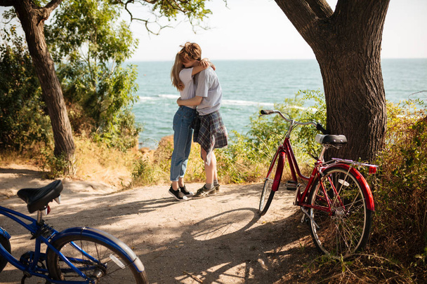 Portret van mooie paar omhelzen elkaar terwijl je in park met geweldig uitzicht op zee op de achtergrond. Jonge leuk paar keer samen met fietsen naast uitgaven - Foto, afbeelding