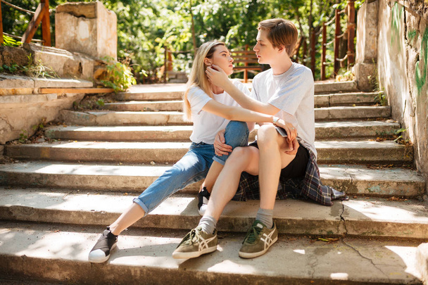 Mooie jonge koppel zittend op een trap in park en omhelzen elkaar terwijl het besteden van tijd samen. Portret van cool jongen en meisje met blond haar zitten en dromerig kijken naar elkaar  - Foto, afbeelding