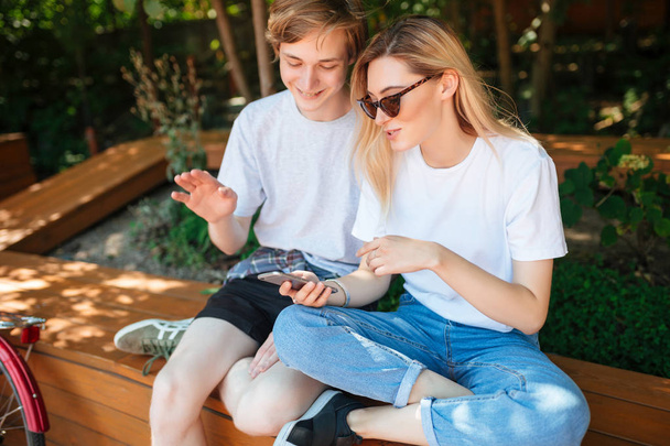 Close up photo of couple sitting on bench in park and happily discussion something while using cellphone. Portrait of young man and pretty lady with blond hair spending time together - Photo, Image