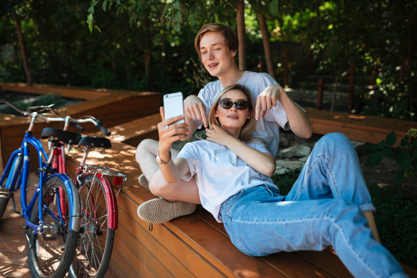 Portret młodej pary, zabawy w parku z rowerów w pobliżu. Chłopiec siedzi na ławce w parku z ładną dziewczyną o blond włosy że opierając się na niego i biorąc selfie Ładna razem na telefon  - Zdjęcie, obraz