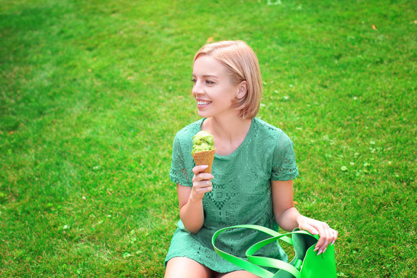 Young happy hipster woman in green lace dress holding ice cream while sitting on grass in park - 写真・画像