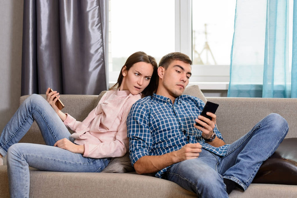 Jong koppel zittend op de Bank en het gebruik van smartphones, verdenkt de vrouw de echtgenoot spionnen op zijn telefoon - Foto, afbeelding