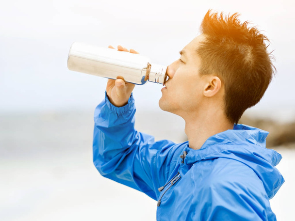 Young man in sport clothes drinking water after workout on the beach - Foto, Imagen