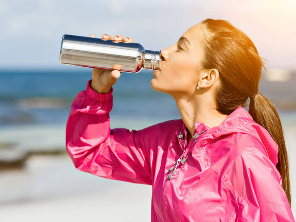 Beautiful girl in sport clothes drinking water after workout on the beach - Foto, Imagen