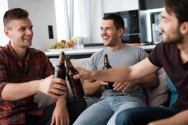 Three men drink beer from dark bottles. They are sitting on the couch and smiling. - Foto, Imagen