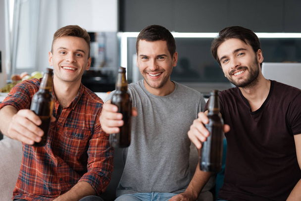 Three men drink beer from dark bottles. They are sitting on the couch and smiling. - Foto, afbeelding