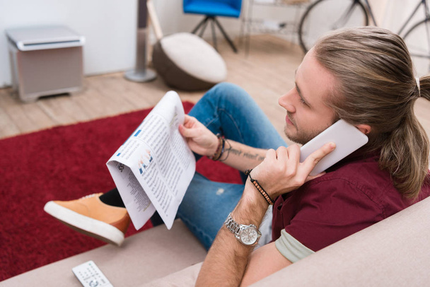 komea mies lukee sanomalehteä ja puhuu älypuhelimella kotona
  - Valokuva, kuva