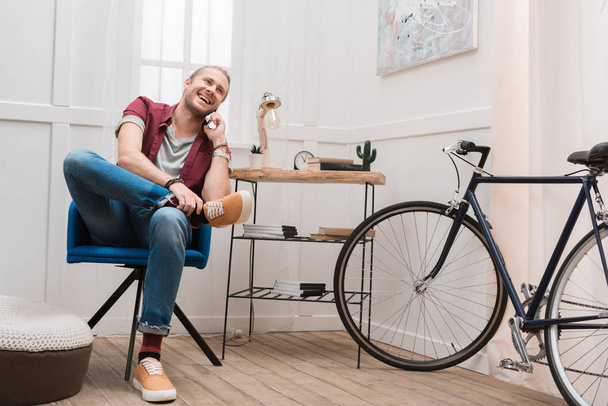bisikleti ile evde sandalyede otururken Smartphone'da konuşan gülümseyen adam - Fotoğraf, Görsel