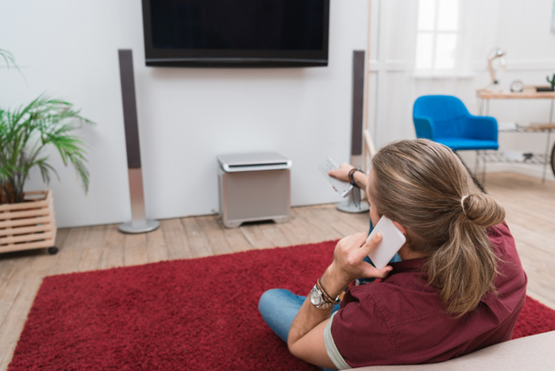 Rückansicht eines Mannes, der mit dem Smartphone spricht und zu Hause fernsieht  - Foto, Bild