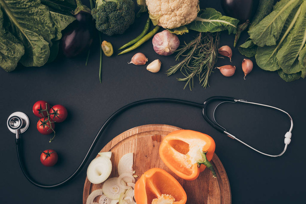 top view of stethoscope between green vegetables and wooden board on gray table - Fotó, kép