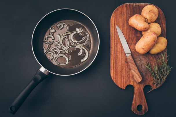 κάτοψη του frying pan με κρεμμύδι και ξύλινη σανίδα με πατάτες στο τραπέζι γκρι - Φωτογραφία, εικόνα