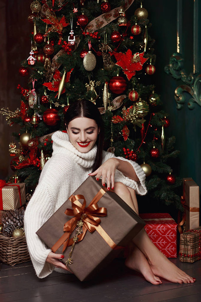 Beautiful girl in sweater sitting at the Christmas tree. Gifts, new year. - Фото, зображення
