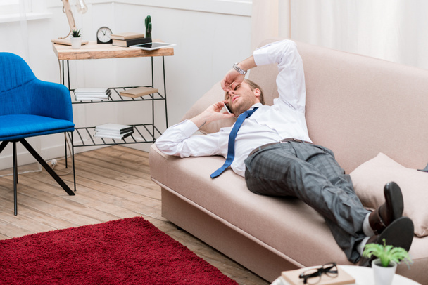 Uomo d'affari stanco sdraiato su un divano a casa e parlando con lo smartphone
 - Foto, immagini