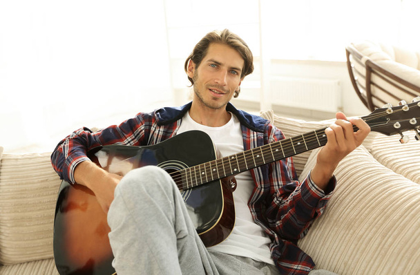 modern guy with guitar sitting on sofa in living room. - Foto, imagen