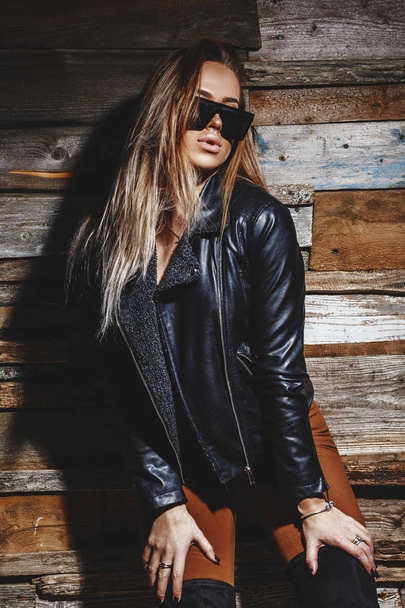Studio shot of young blonde girl. Beauty woman in stylish bra, jacket and sunglasses standing on grunge wooden wall background  - Foto, afbeelding
