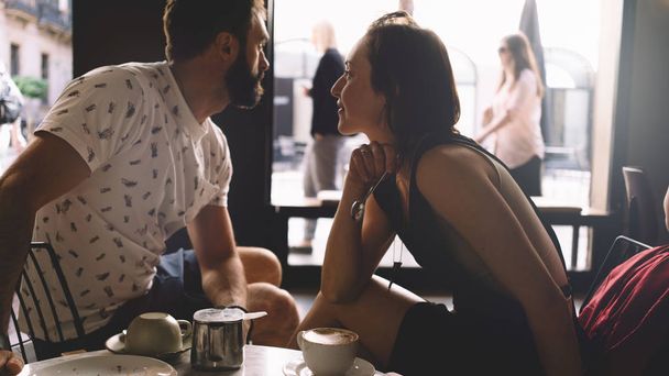 Couple of young beautiful people is sitting in a modern coffee shop on a summer day. A romantic dating of a handsome bearded guy and a charming girl in a cafe. Portrait of a man and a woman. - 写真・画像