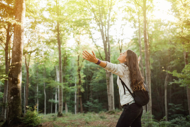 Young woman emgracing the sun and nature with backpack in the forest on sunset light in the autumn season, looking up, with her hands in the air, exploring the nature. - 写真・画像
