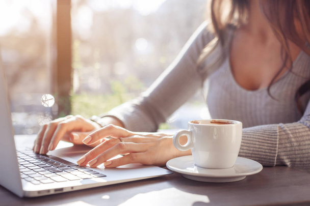 A girl works behind a laptop in a cafe, fingers typing on the keyboard. Workplace in a cafe, a cup of coffee next to a laptop, toned bright sunny photo - Foto, imagen