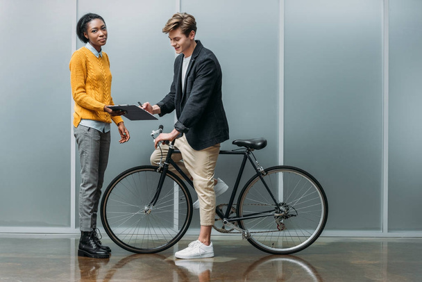 jonge zakenman ondertekening contract in handen van jonge Afro-Amerikaanse collega terwijl hij zittend op de fiets - Foto, afbeelding