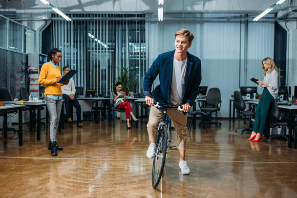 onnellinen nuori liikemies ratsastus pyörä moderni toimisto, kun hänen kollegansa työskentelevät
 - Valokuva, kuva
