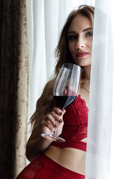 Sensual young woman dressed in a red lingerie  drinks  wine  nea a window. - Фото, изображение