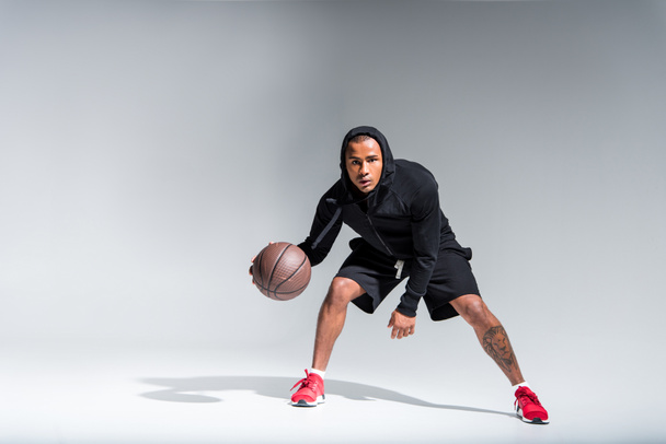 deportivo afroamericano hombre jugando con pelota de baloncesto y mirando a la cámara en gris
  - Foto, Imagen